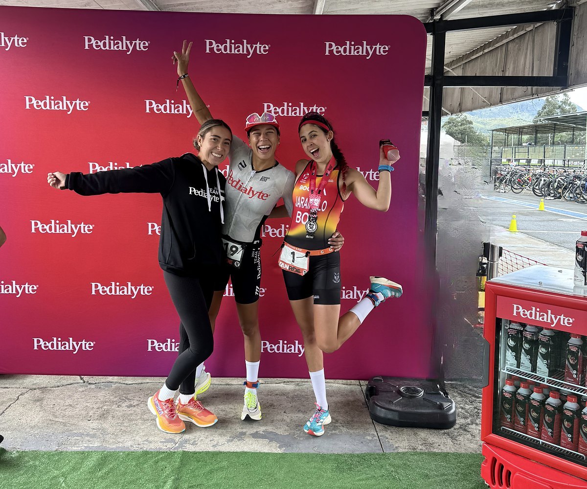 María and two team athletes jumping and cheering in front of a sponsor backdrop