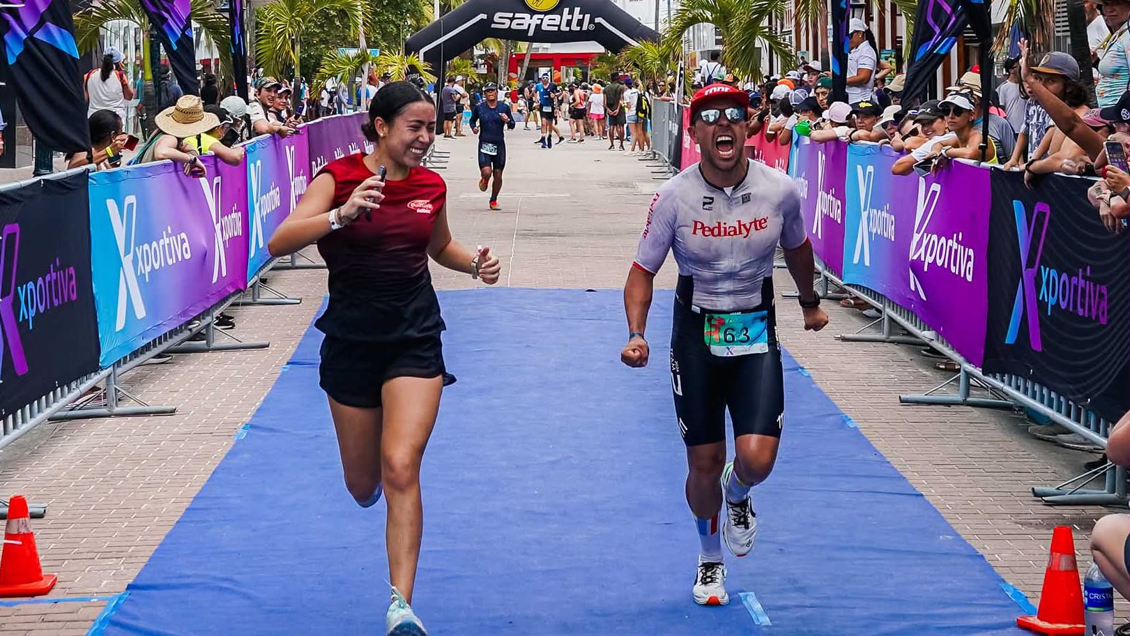 María running beside a team athlete as he crosses the finish line at Tri Fest San Andrés, both smiling