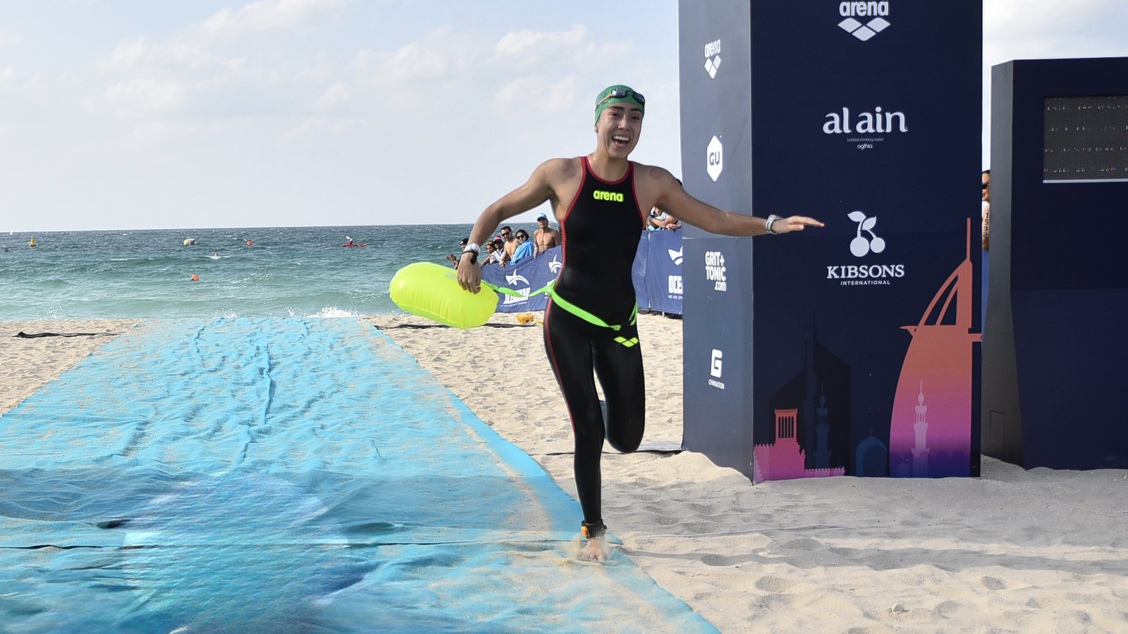 María running up the beach to the finish of the Oceanman World Final in Dubai, arms open, with her yellow tow buoy, the sea behind her